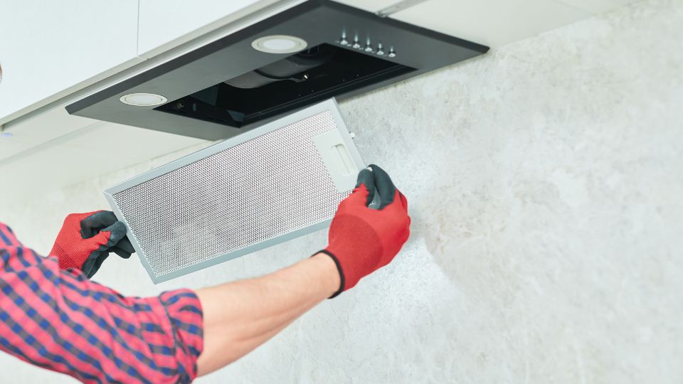 Person wearing red gloves is removing a metal filter from a kitchen range hood at a house cleaning session. The individual is dressed in a red and blue plaid shirt, and the background features a light-colored wall.