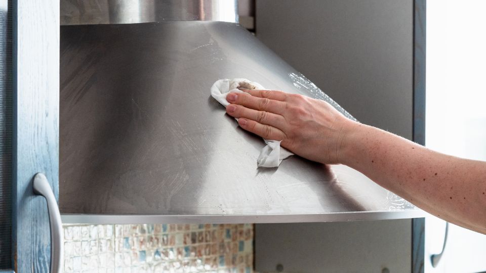 A person from Scottsdale house cleaning is meticulously wiping a stainless steel kitchen hood with a cloth. The hand moves in a circular motion on the metallic surface, while part of the tile backsplash peeks into view, showcasing professional cleaning services at work.