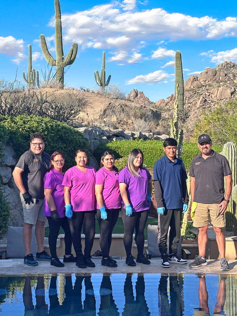Team 1 of Fernandez Exclusive Services of seven people stands outdoors in front of a pool, with desert scenery and cacti in the background. They are smiling, with most wearing casual clothing and purple shirts. The sky is blue with a few clouds.