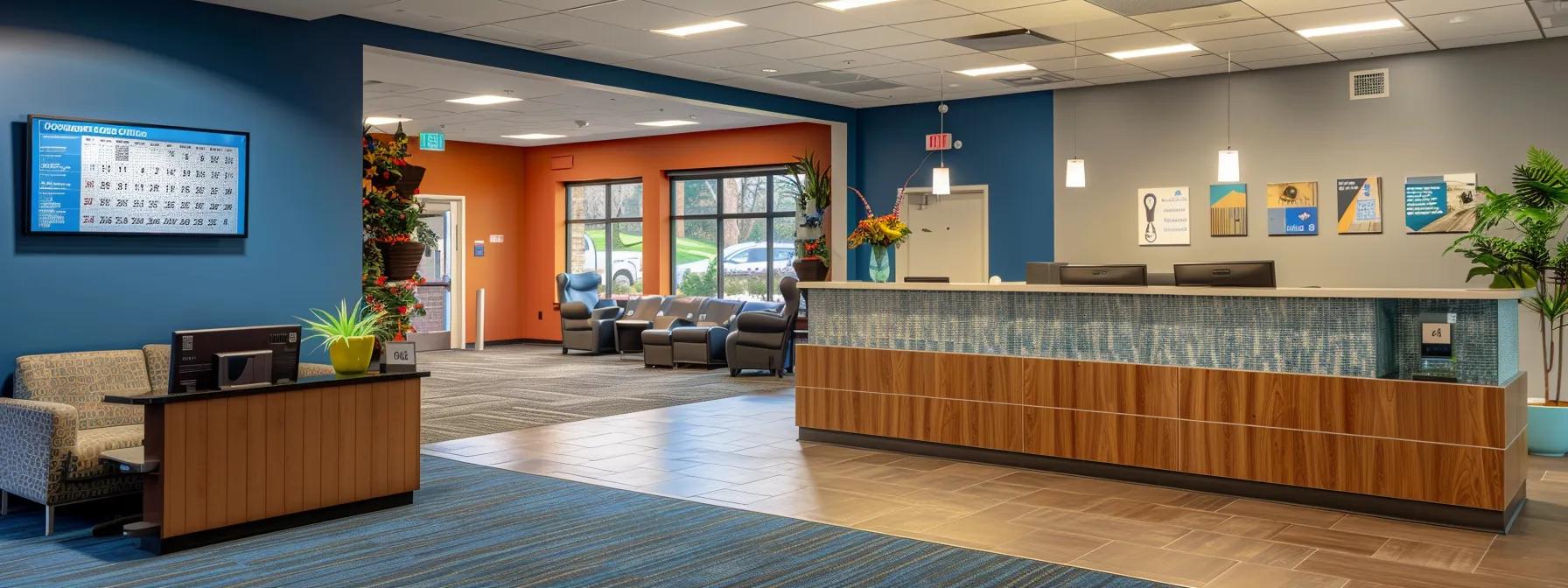 A modern clinic reception area with a wooden front desk, blue accent walls, comfortable seating, wall art, potted plants, and large windows letting in natural light. A digital information board is visible on the wall.