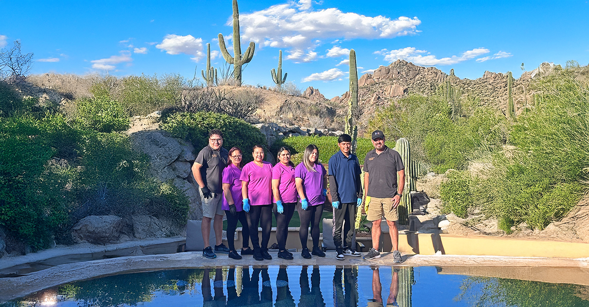 Seven people stand beside a pool in a desert landscape with cacti and rocky hills. Six wear matching purple shirts and black pants, while one on each end wears casual clothing. The sky is bright with scattered clouds.