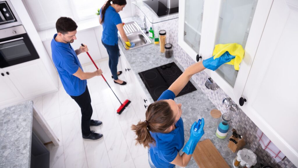 Professional cleaning team in blue shirts cleaning a modern kitchen, one person wiping cabinets, another using a broom, showcasing expert house cleaning services in Scottsdale.