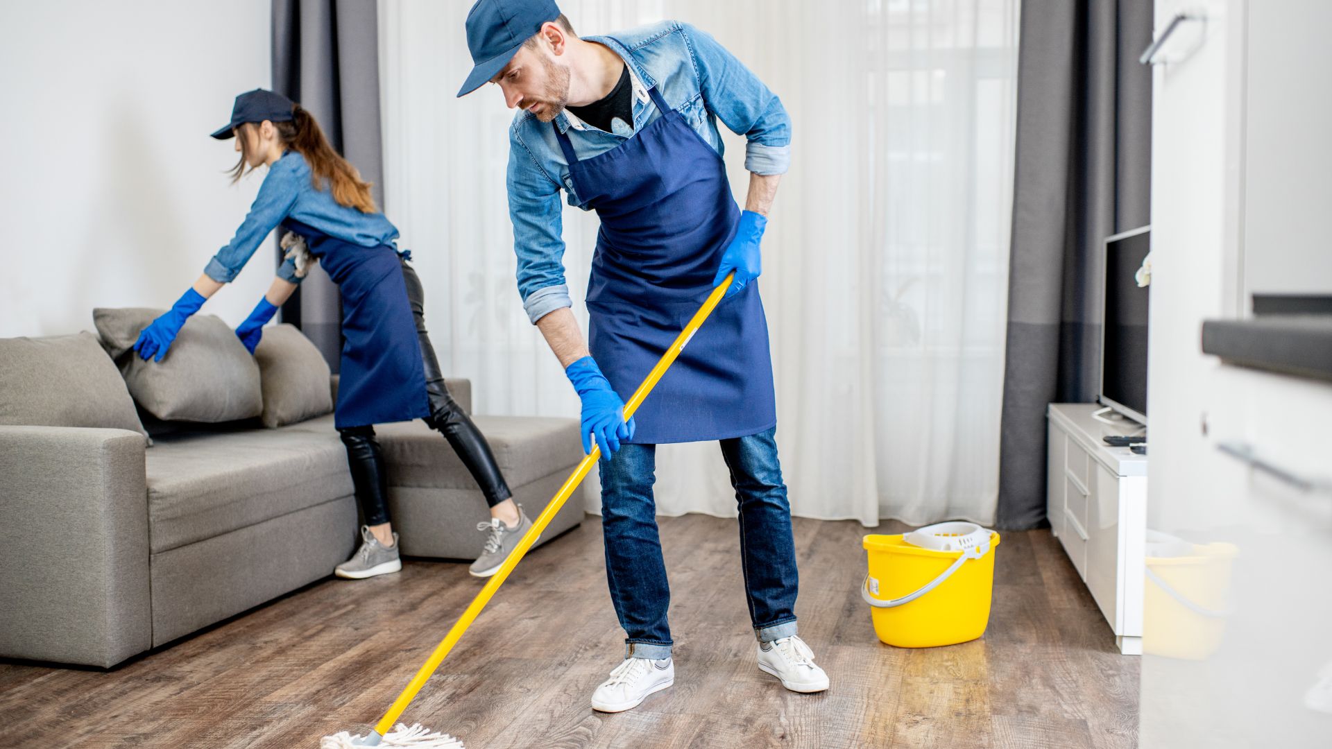 Professional cleaners mopping floor and organizing cushions in modern living room, showcasing house cleaning services.